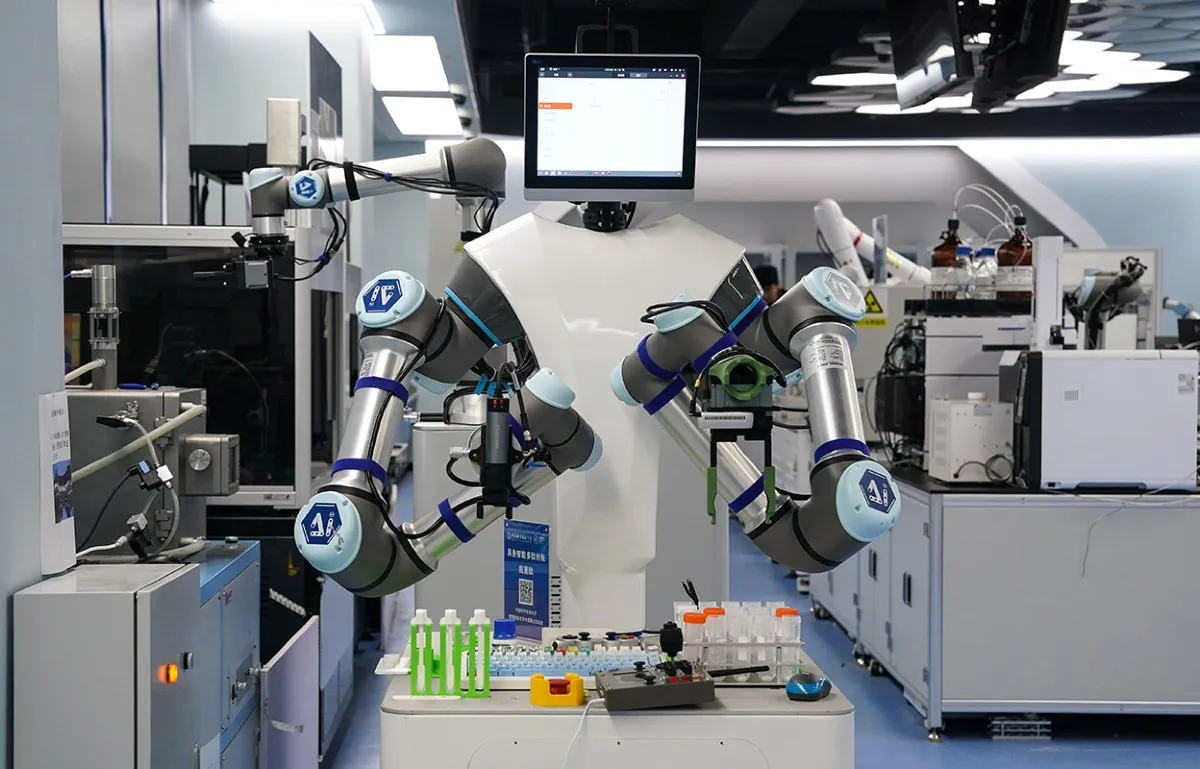 Robô de laboratório com dois braços articulados e uma tela no lugar da cabeça, manipulando tubos e racks de amostras numa bancada cercada de instrumentos