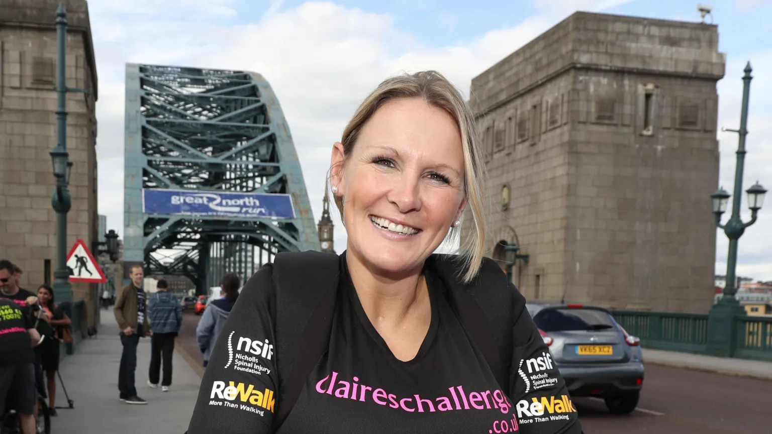 Claire Lomas sorrindo em frente à Ponte Tyne, usando camiseta do projeto ReWalk