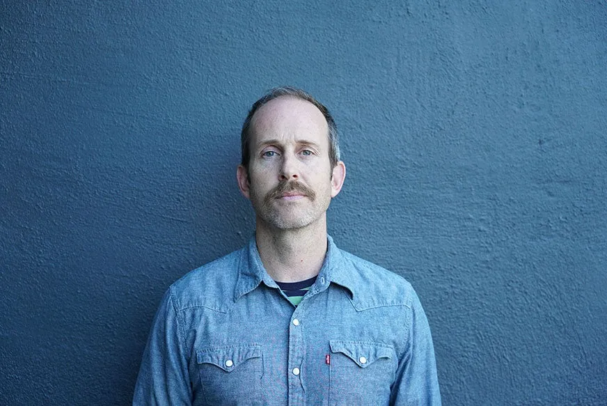 Bruce Straley, de camisa jeans e bigode, encostado numa parede azul olhando para a câmera