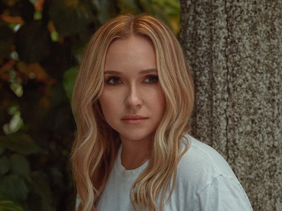 Retrato de Hayden Panettiere, de cabelo loiro e camiseta branca, encostada em uma árvore