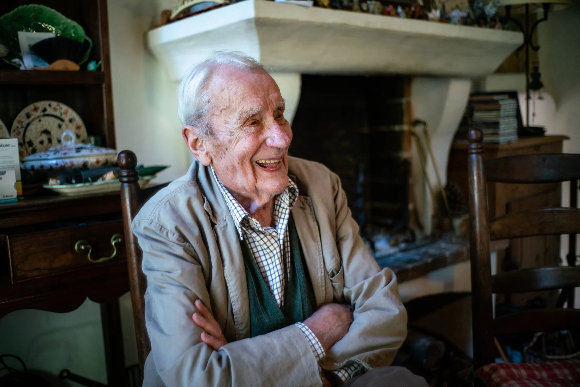 Christopher Tolkien sentado em cadeira de madeira com braços cruzados e sorrindo, diante de uma lareira em ambiente doméstico