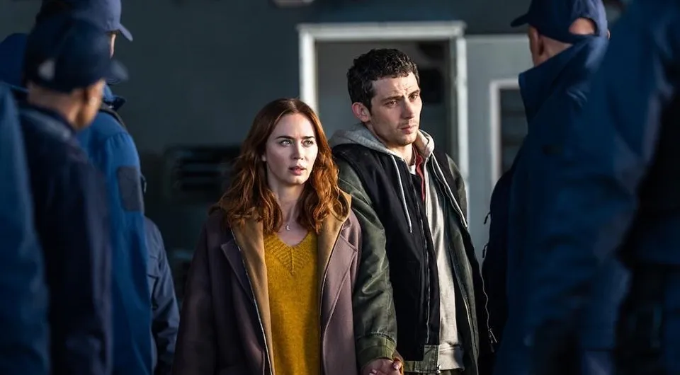 Emily Blunt e Josh O'Connor de mãos dadas cercados por homens de uniforme em cena de Dia D