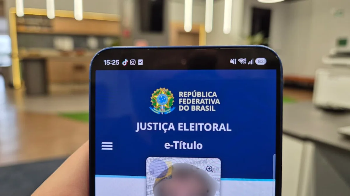 Mão segurando um celular com o aplicativo oficial e-Título da Justiça Eleitoral aberto, com a foto do título de eleitor borrada