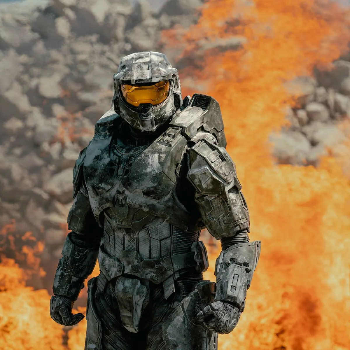 Master Chief de armadura completa e capacete de visor dourado, caminhando à frente de uma parede de fogo e fumaça