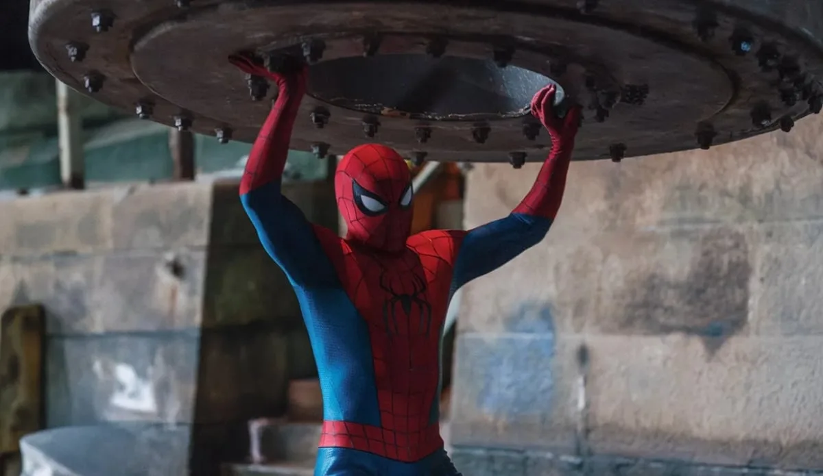 Homem-Aranha, de uniforme vermelho e azul, ergue uma enorme estrutura circular de metal acima da cabeça em cena de Um Novo Dia
