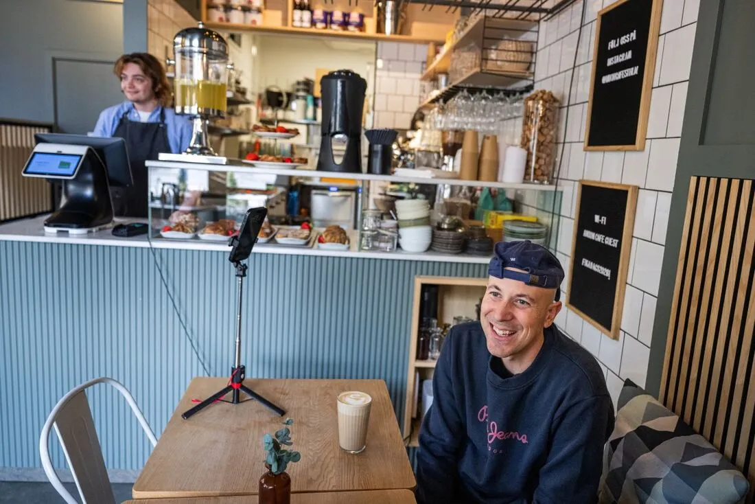 Homem sorrindo em mesa do Café Mona em Estocolmo com celular em suporte e latte; barista ao fundo atrás do balcão