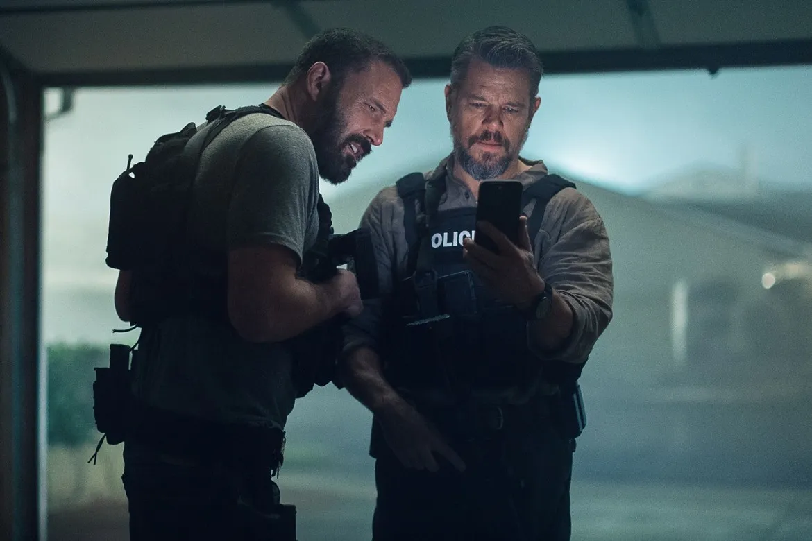 Ben Affleck e Matt Damon, de colete policial, olham juntos para a tela de um celular em cena de Dinheiro Suspeito