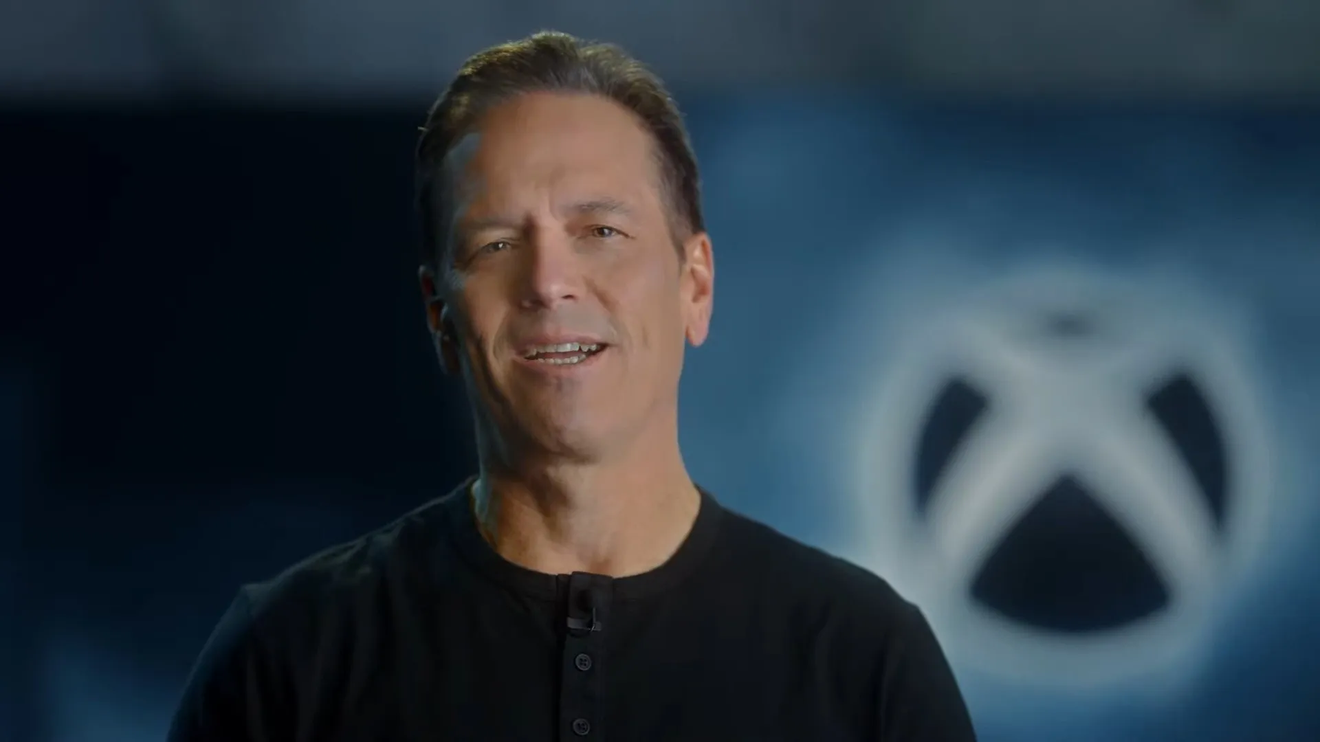Phil Spencer sorrindo em frente ao logo do Xbox durante evento da Microsoft