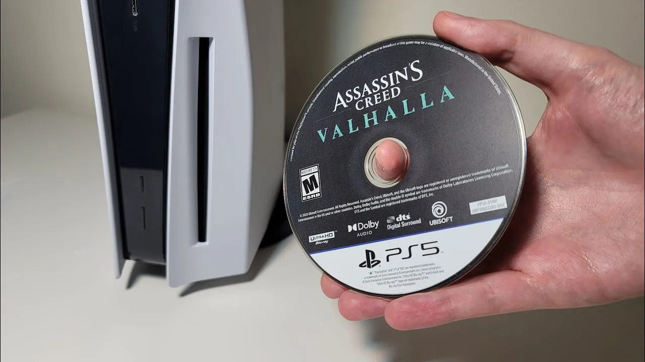 Mão segurando o disco de Assassin's Creed Valhalla para PS5 em frente ao console PlayStation 5, simbolizando o fim da mídia física anunciado pela Sony para 2028