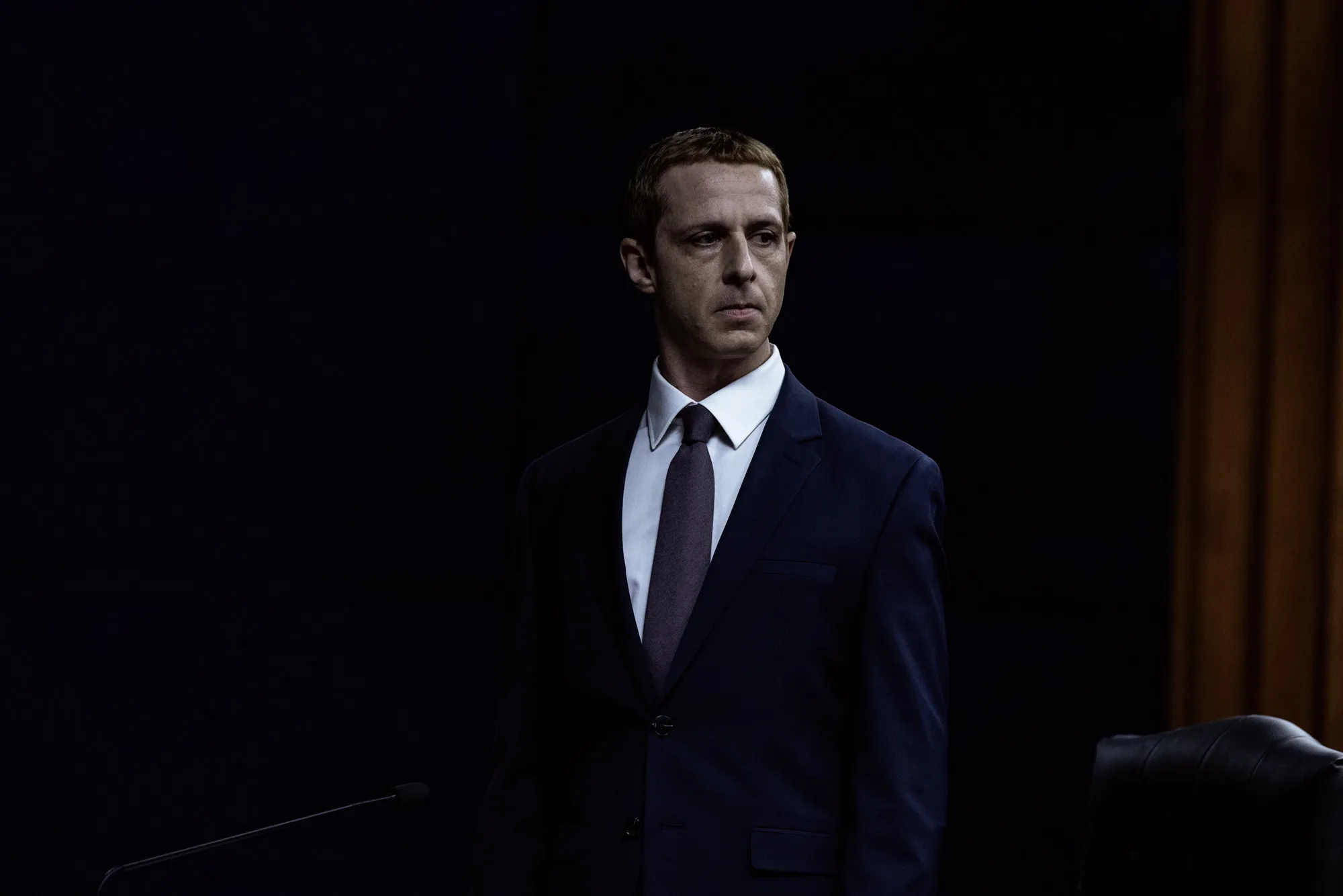 Jeremy Strong como Mark Zuckerberg, de terno escuro e gravata, de pé diante de um microfone em sala de audiência quase tomada pelo breu