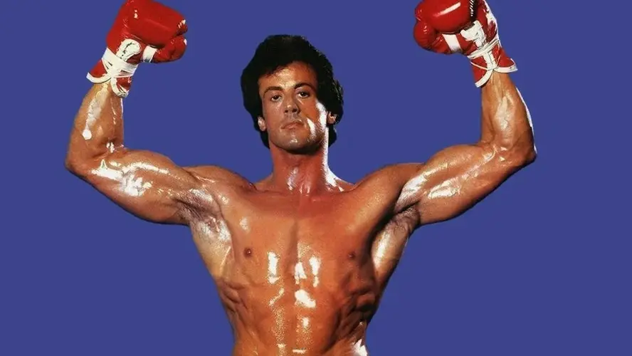 Sylvester Stallone como Rocky Balboa, sem camisa e com o corpo definido, erguendo os braços com luvas de boxe vermelhas diante de um fundo azul em foto de divulgação de Rocky III: O Desafio Supremo