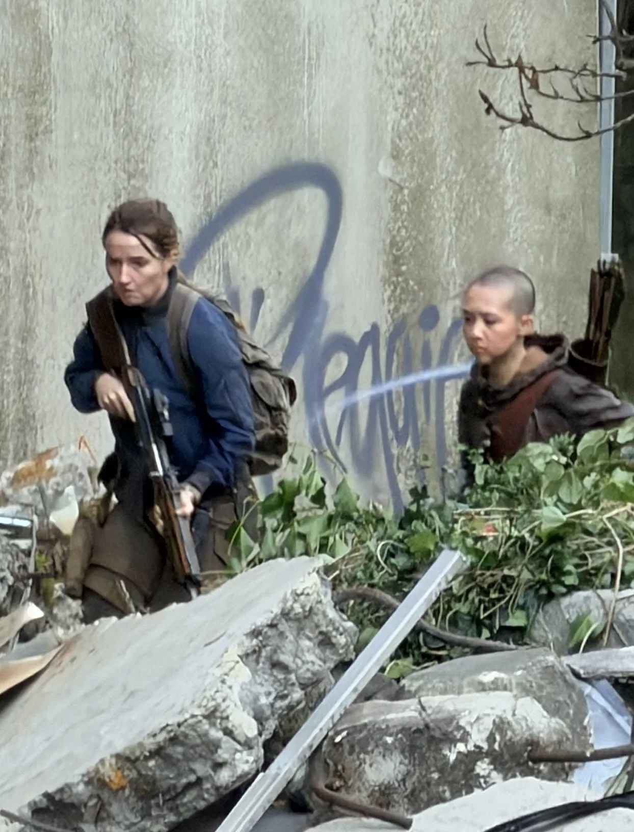 Vista mais próxima de Abby com rifle e Lev com coldre de flechas, parede de concreto com grafite azul ao fundo